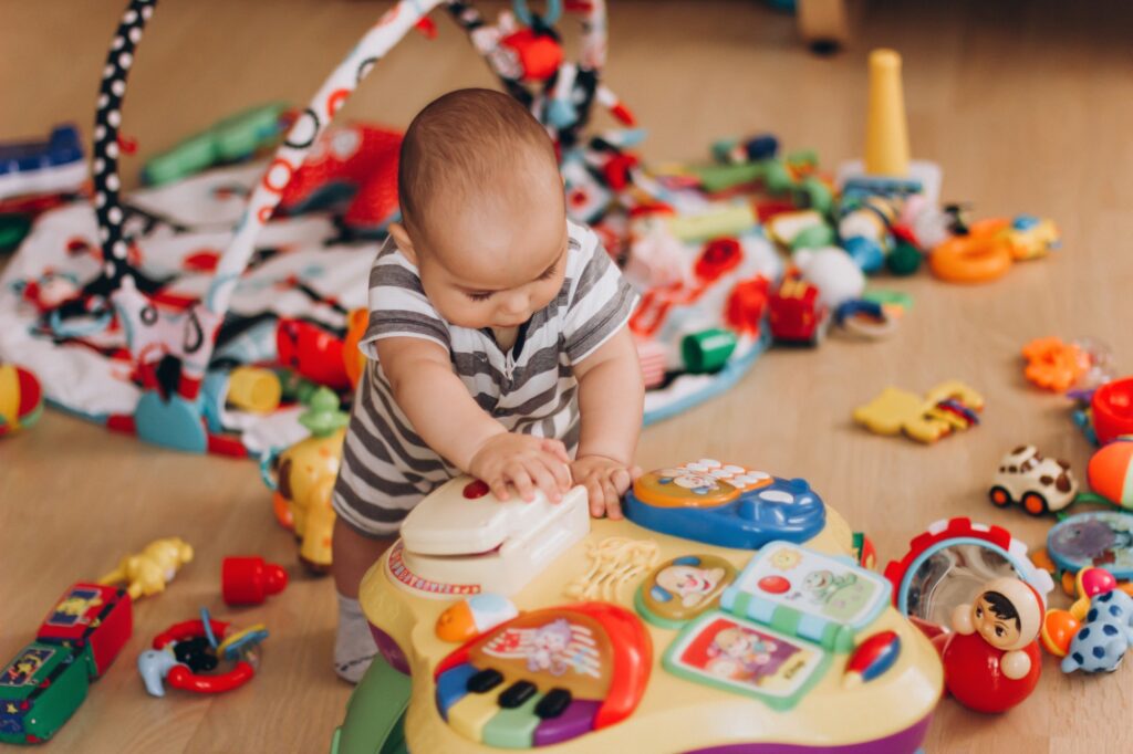 Cute chubby baby stands and plays with a children's music table. Lots of colorful toys everywhere.