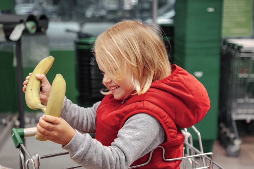 Cute toddler little girl sitting in shopping cart in store or market and buying goods on store shelf